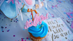 A cupcake with blue icing with a candle that says "It's a girl" sitting on a table.