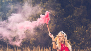 Woman posing in a field holding a smoke stick emitting pink smoke.