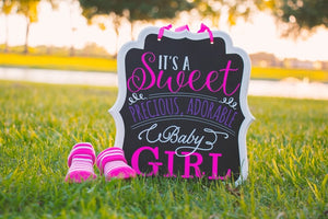 Chalk sign with "It's a sweet precious, adorable baby girl" written on it, sitting in grass next to pink slippers.