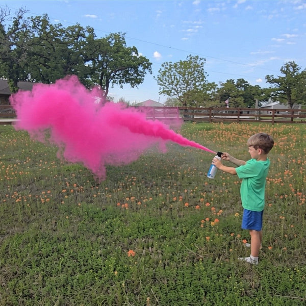 Child holding a pink smoke device in a grassy field with trees and a building in the background